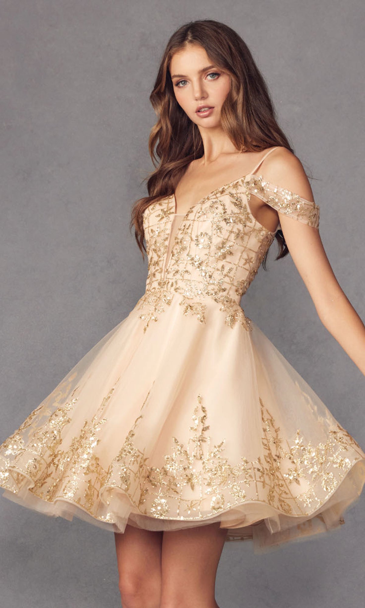 A woman models the Glitter-Print Short Babydoll Homecoming Dress 903, a short off-shoulder champagne dress with gold floral embroidery, standing against a plain gray background.